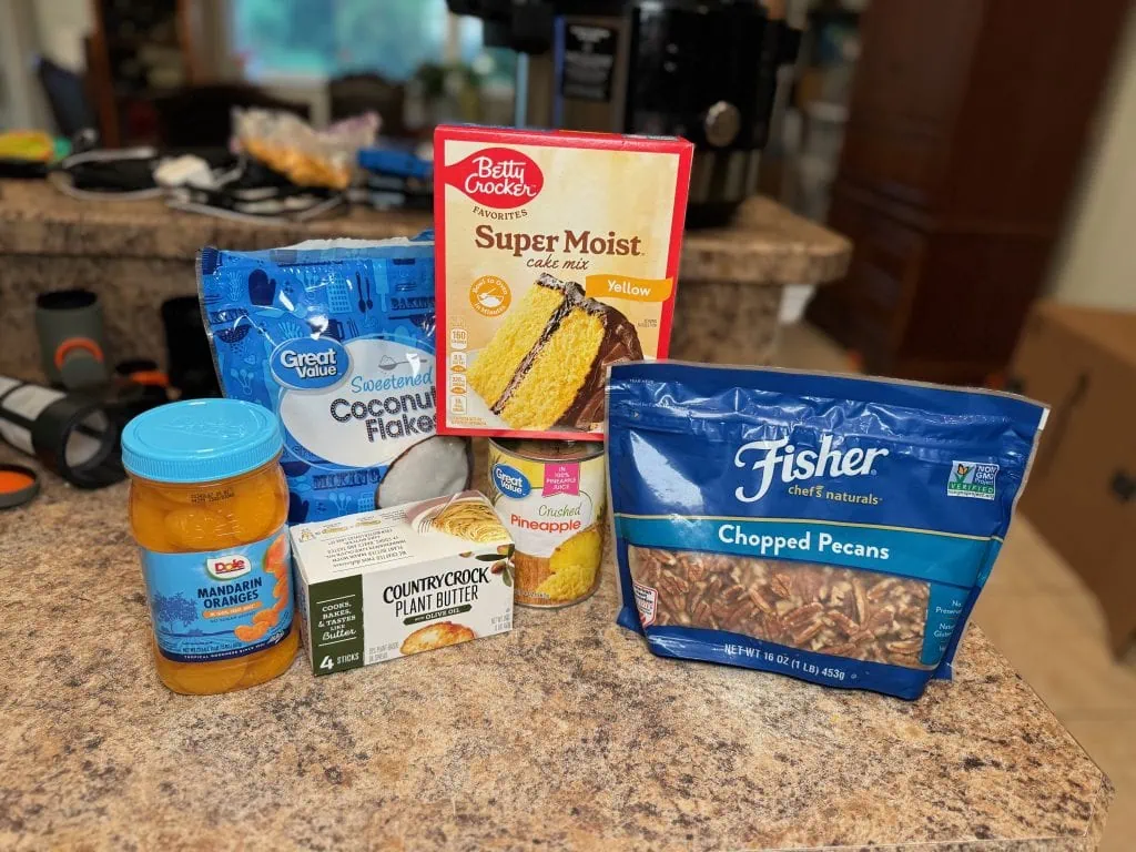 Ingredients for a dairy-free Hawaiian dump cake displayed on a kitchen counter, including yellow cake mix, crushed pineapple, mandarin oranges, shredded coconut, chopped pecans, and plant-based butter.